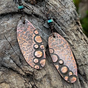 May include: A pair of handcrafted copper earrings. Each earring features an oval shape with a textured surface and a series of embossed circles along one side. The earrings have a dark patina and are accented with small turquoise beads.