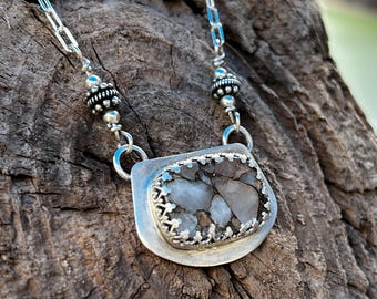 May include: A silver necklace with a rectangular pendant featuring a mosaic of gray and brown stones. The pendant is framed with a decorative silver border and hangs from a delicate chain with beaded accents.