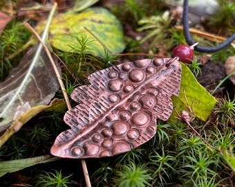 May include: A copper-colored leaf pendant with a textured surface and a black cord necklace. The pendant has a detailed design with raised circles and lines. The background features green moss, leaves, and a small red bead.