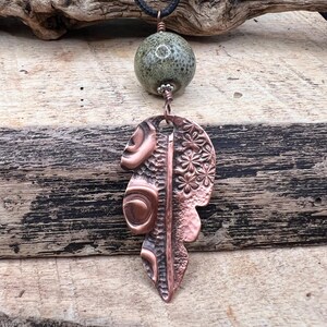 May include: A handcrafted necklace featuring a green speckled stone bead and a copper leaf pendant. The leaf pendant has intricate embossed designs and is suspended from a black cord. The necklace is displayed against a rustic wooden background.