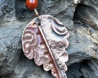 May include: A handcrafted copper pendant necklace featuring a leaf-shaped design with intricate embossed details. The pendant is suspended from a black cord and accented with a small, round, reddish-orange bead. The necklace is a unique piece of jewelry.