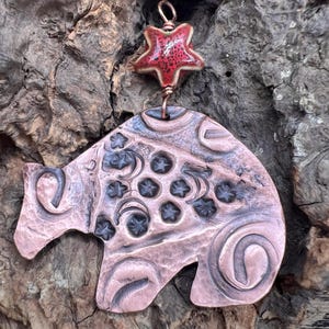 May include: Copper pendant in the shape of a bear with etched details and star accents. A red star-shaped bead hangs above the bear pendant. The pendant is suspended by a copper wire.