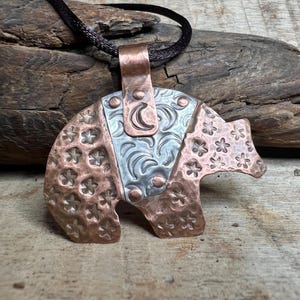 May include: A handcrafted copper bear pendant with a silver-colored center section. The bear's body is adorned with floral patterns and a crescent moon design. The pendant hangs from a black cord, showcasing a rustic, artistic style.