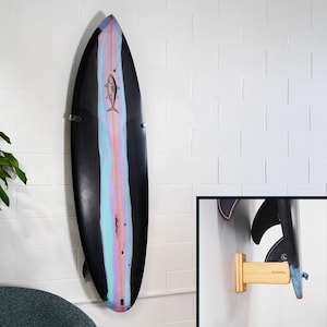May include: A black surfboard with a colorful vertical stripe in shades of pink, blue, and purple. A fish graphic is centered on the board. The surfboard is mounted on a white brick wall with metal brackets.