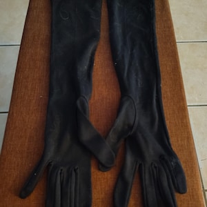 Peut inclure: Paire de longs gants noirs, probablement pour une tenue de soirée, posés sur une surface marron. Les gants s'étendent au-delà du poignet, couvrant une grande partie du bras. Le matériau semble être un tissu doux.