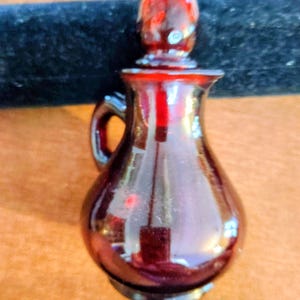 Può includere: Una piccola ampolla o caraffa in vetro rosso intenso con corpo arrotondato, manico curvo sul lato e un tappo in vetro appuntito abbinato. Il vetro presenta una superficie liscia e riflettente con una qualità traslucida che cattura la luce.
