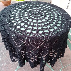 Peut inclure: Une nappe ronde en crochet noir présentant un motif ajouré complexe avec un design central en étoile qui rayonne vers l'extérieur en formes géométriques répétitives et un bord festonné qui retombe sur les côtés d'une table circulaire.