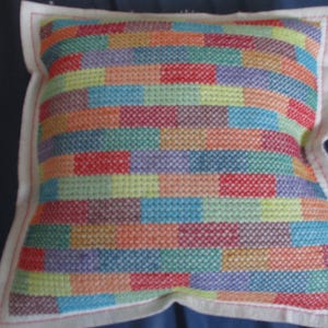 Puede incluir: Una almohada colorida con un patrón geométrico de bloques rectangulares en tonos de rojo, naranja, amarillo, verde, azul, morado y marrón. La almohada está hecha de una tela blanca con un borde rojo.