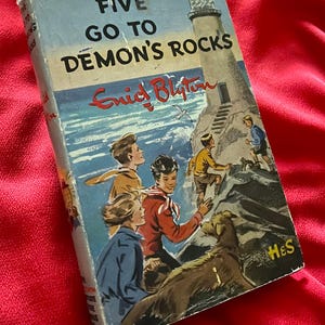以下が含まれることがあります： エニッド・ブライトン著「Five Go to Demon's Rocks」のヴィンテージ本。表紙には、灯台の近くで探検する子供たちと犬のイラストが描かれています。タイトルと著者の名前が目立つように表示されています。