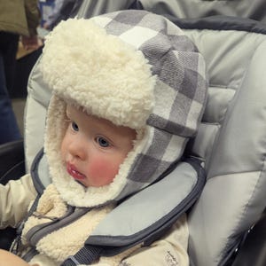 May include: A baby wearing a gray and white plaid trapper hat with a fluffy, cream-colored trim. The baby is in a stroller and wearing a cream-colored onesie. The stroller has gray padding.