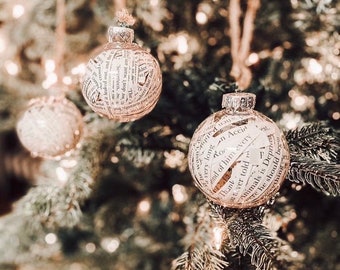 May include: Two glass Christmas ornaments decorated with white paper scraps with text printed on them. The ornaments are hanging on a green Christmas tree.