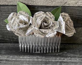 Puede incluir: Un peine para el cabello con dos rosas blancas de papel unidas. Las rosas están hechas de páginas de un libro y tienen hojas verdes. El peine es plateado y tiene una fila de dientes.