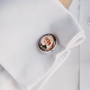 May include: Close-up of a white dress shirt with French cuffs. The shirt features a pair of round, silver cufflinks with a floral design in shades of pink, orange, and green. The cufflinks are the focal point.