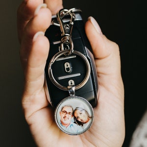 May include: A black car key fob with a silver keychain and a round pendant. The pendant features a color photograph of two people. The key fob is held in a hand.