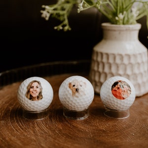May include: Three white golf balls with personalized photos on them. The photos are of a woman, a dog, and a couple. The golf balls are on a wooden surface.