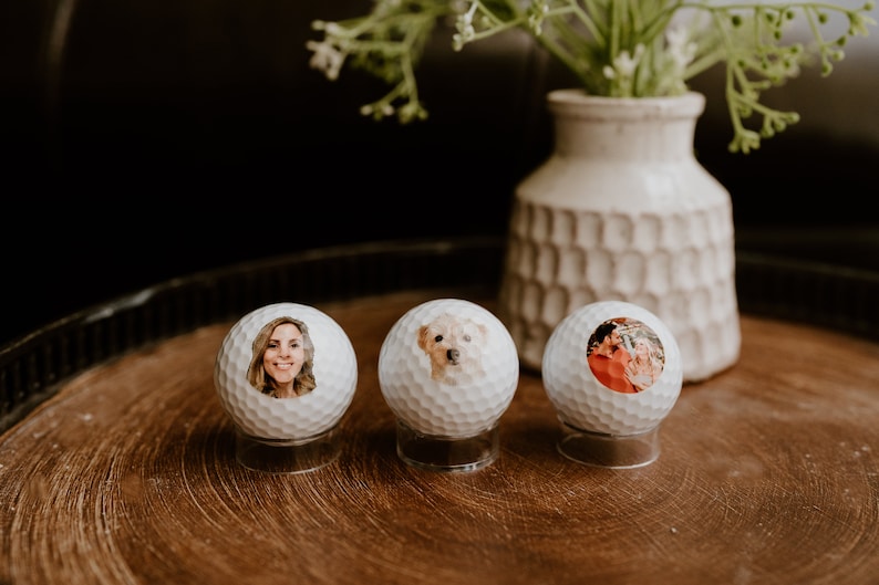 May include: Three white golf balls with personalized photos on them. The photos are of a woman, a dog, and a couple. The golf balls are on a wooden surface.