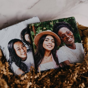 May include: Three friends are pictured on two slate coasters. The coasters are in a brown wicker basket with brown shredded paper.