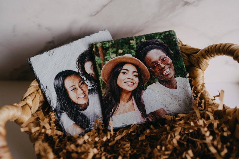 May include: Three friends are pictured on two slate coasters. The coasters are in a brown wicker basket with brown shredded paper.