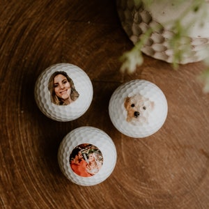 May include: Three white golf balls with personalized photos printed on them. The photos include a woman, a dog, and a couple.