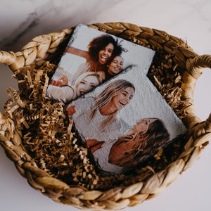 May include: Two square photo coasters with images of friends smiling and laughing. The coasters are in a woven basket filled with brown packing paper.
