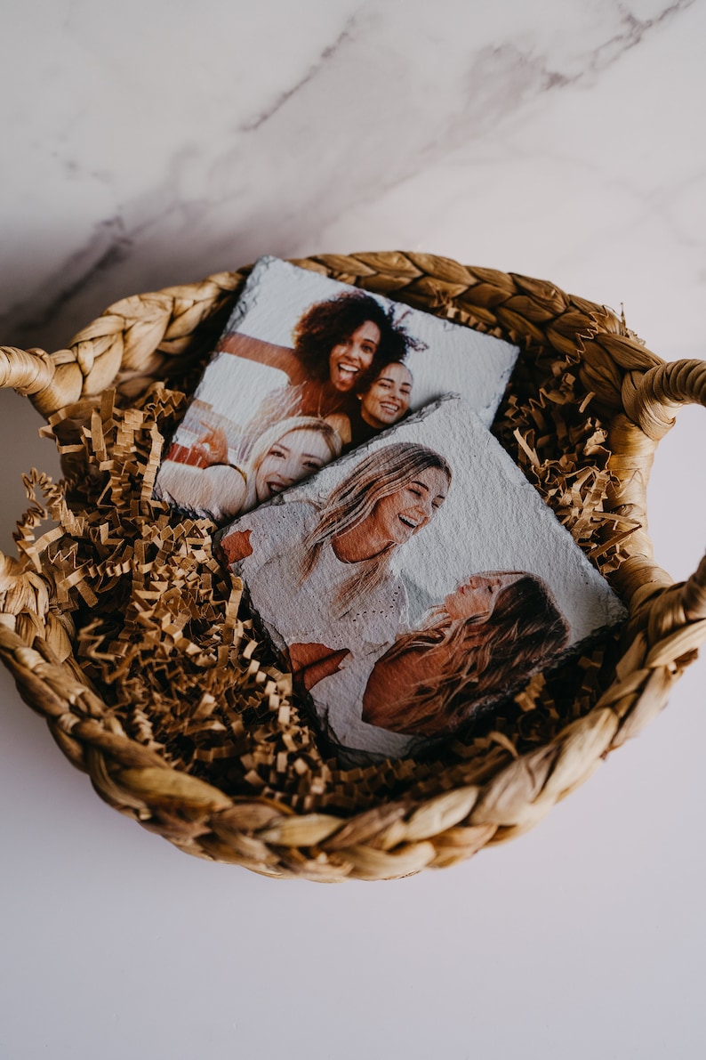 May include: Two square photo coasters with images of friends smiling and laughing. The coasters are in a woven basket filled with brown packing paper.