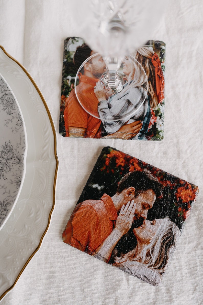 May include: Two square slate coasters with a printed photo of a couple kissing. The coasters are on a white tablecloth.