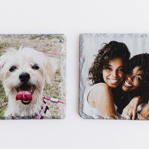 May include: Two square slate coasters with images printed on them. One coaster features a white dog with pink tongue sticking out. The other coaster features two women hugging each other.