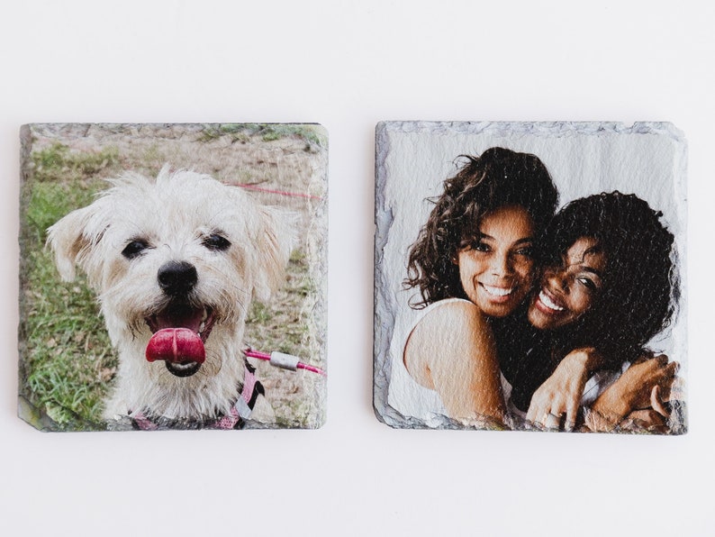 May include: Two square slate coasters with images printed on them. One coaster features a white dog with pink tongue sticking out. The other coaster features two women hugging each other.