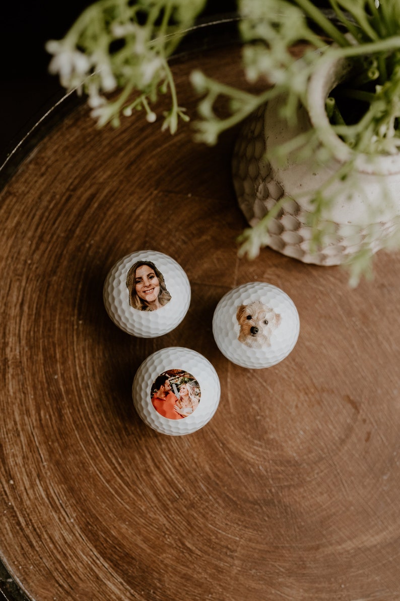 May include: Three white golf balls with personalized photos printed on them. The photos include a woman, a couple, and a dog.