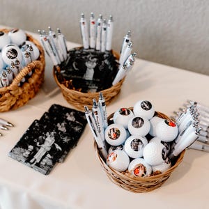 May include: Three wicker baskets filled with white golf balls with personalized photos and white pens. The baskets are on a white tablecloth.