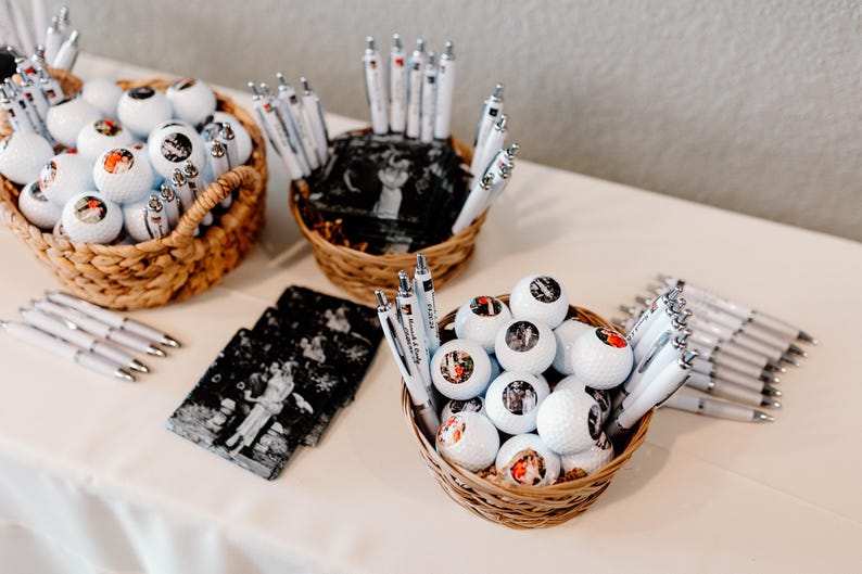 May include: Three wicker baskets filled with white golf balls with personalized photos and white pens. The baskets are on a white tablecloth.