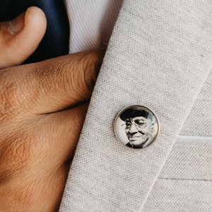 May include: A silver lapel pin with a black and white photo of a man wearing a hat and glasses. The pin is on a gray blazer.