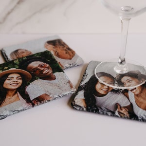 May include: Four square slate coasters with photos of friends printed on them. The coasters are arranged in a square shape with a wine glass on top of one of the coasters.