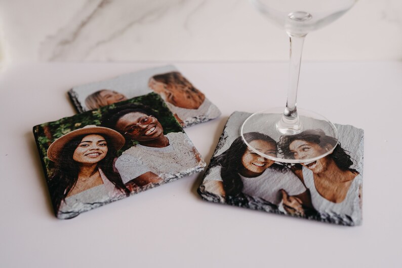 May include: Four square slate coasters with photos of friends printed on them. The coasters are arranged in a square shape with a wine glass on top of one of the coasters.