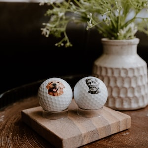 May include: Two white golf balls with personalized images on them. One ball has a picture of a golden retriever, and the other has a picture of an older man. The golf balls are sitting on a small wooden stand.