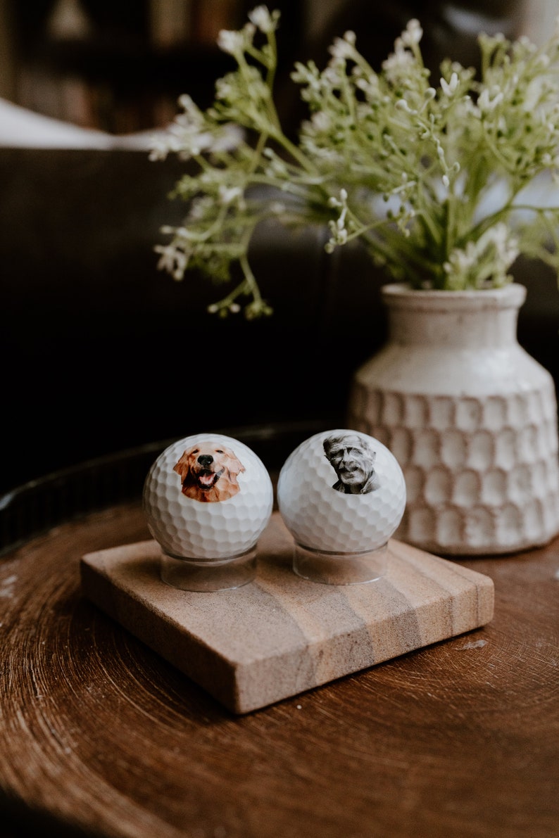 May include: Two white golf balls with personalized images on them. One ball has a picture of a golden retriever, and the other has a picture of an older man. The golf balls are sitting on a small wooden stand.
