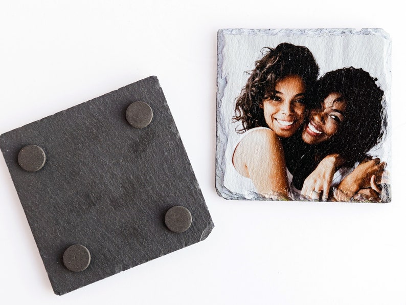 May include: Two women are smiling and hugging in a photo printed on a black slate coaster. The coaster has four cork feet on the bottom.