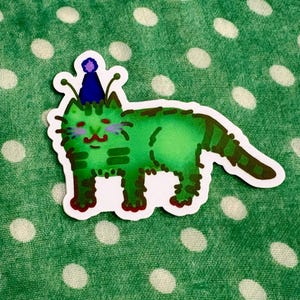 May include: A green cat sticker with a party hat. The cat has a smiling face, purple eyebrows, and brown stripes. The sticker is on a green fabric with white polka dots.
