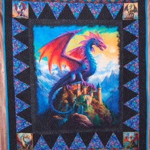 May include: A colorful quilt featuring a fantasy dragon perched atop a castle on a mountain. The dragon has purple and blue scales with red and orange wings. The quilt has a black border with blue and purple triangle accents and a blue border.