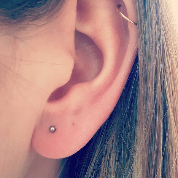 May include: Close-up of a person's ear with two piercings. The lower piercing has a small silver stud, and the upper piercing has a gold hoop.