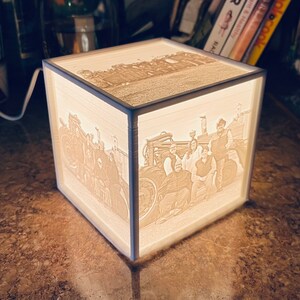 May include: A white cube-shaped lamp with a 3D image of a group of people standing in front of a tractor. The image is illuminated from behind, creating a soft glow.
