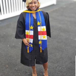 May include: A person wearing a black graduation gown and cap, with a blue, red, and white sash with yellow stars. The sash is a representation of the flag of Curaçao.