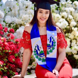 May include: A woman wearing a red dress and a blue and white graduation sash with the flag of El Salvador. She is wearing a black graduation cap and smiling. The background is a white and red flower bush.