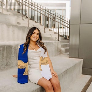 May include: A blue and white graduation stole with a gold eagle design and gold stars. The stole is draped over a white dress. The person is sitting on concrete steps, smiling at the camera.