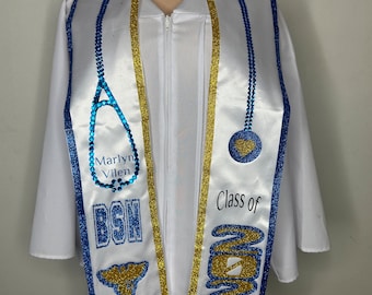 Op de afbeelding: Witte afstudeerjurk met een blauwe en gouden afstudeersjerp. De sjerp heeft een stethoscoop, een hart, de tekst "Marlyn Vilen", "BSN" en "Klas van 2024" in blauw en gouden glitter.