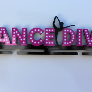 May include: A black medal display with the words "DANCE DIVA" in pink letters with white polka dots. A silhouette of a dancer is in the center of the display.