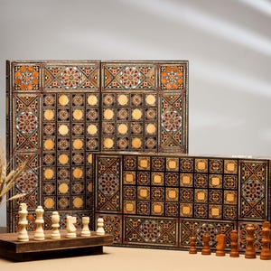 Puede incluir: Un tablero de juego de madera con intrincados diseños de incrustaciones en tonos marrones, dorados y negros. El tablero está dividido en cuadrados y círculos, lo que sugiere que es para jugar a varios juegos. También hay piezas de ajedrez en un tablero de madera más pequeño en primer plano.