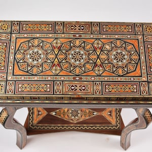 May include: An intricately designed wooden table with a mosaic top. The tabletop features geometric patterns in shades of brown, orange, and black. The legs are curved and the table has a lower shelf with matching mosaic details. A unique piece of furniture.