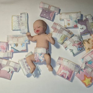 以下が含まれることがあります： ピンク、ブルー、イエローなど、さまざまな色とパターンでミニチュアのベビーおむつが揃っています。おむつは、星、花、動物など、さまざまなデザインが施されています。小さなリアルなベビー人形がおむつの上に横たわっています。