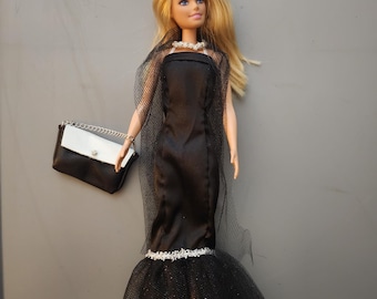May include: A blonde doll wearing a black strapless gown with a tulle overlay and a white beaded trim. The doll is holding a black and white purse.